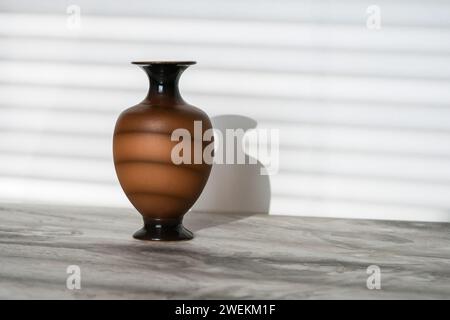 Vaso marrone su un piano in marmo grigio vicino a una parete bianca illuminata da raggi solari attraverso le tende Foto Stock