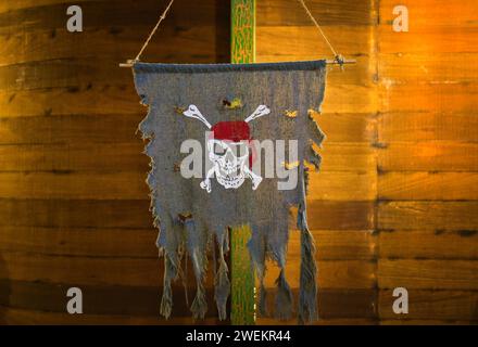 Striscione pirata teschio e ossa, noto anche come bandiera Jolly Roger, appeso allo scafo di una nave di legno. Concetto di pirateria, furto. Foto Stock