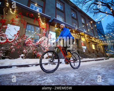 Un uomo cavalca una bicicletta lungo una strada splendidamente decorata in una città invernale. Uomo barbuto con una giacca blu su una bicicletta rossa. Città a Natale Foto Stock