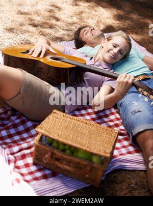 Coppia felice, picnic e chitarra per amore, romanticismo o musica all'aperto, divertimento o relax insieme nella natura. Uomo e donna sorridono con Foto Stock