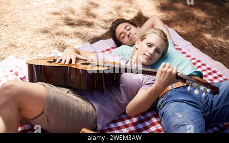 Coppia, picnic e chitarra per romanticismo, musica o amore all'aperto, divertimento o relax insieme nella natura. Giovane uomo e donna sorridono con Foto Stock