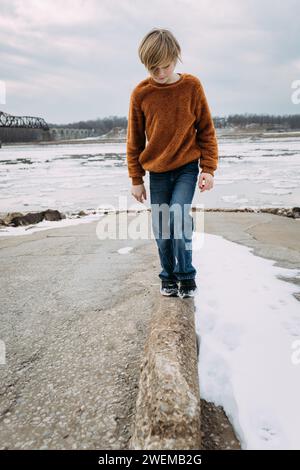 Un bambino pre-adolescente che cammina lungo la riva del fiume nelle nevose giornate invernali Foto Stock