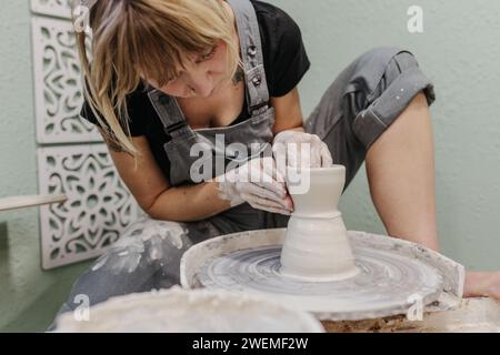 La donna siede alla ruota della ceramica mentre tira pentole in studio domestico Foto Stock