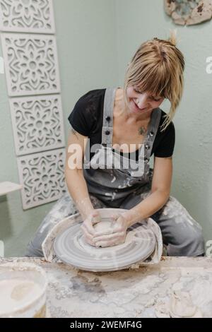 La donna siede alla ruota della ceramica mentre tira pentole in studio domestico Foto Stock