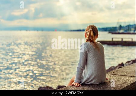 Graziosa bambina che riposa vicino al lago, ammirando un tramonto incredibile, raggi di luce che cadono tra le nuvole Foto Stock