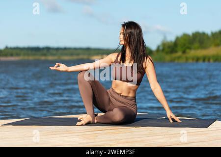 Una donna attraente che pratica yoga si rilassa dopo un allenamento seduto sulla riva di un lago, mostra il simbolo del mudra, si allena con leggings e A. Foto Stock