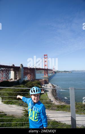 Ragazzo che indossa un casco da bicicletta che punta al ponte sospeso Golden Gate che attraversa il Golden Gate in una giornata di sole con un ponte alle sue spalle. Foto Stock