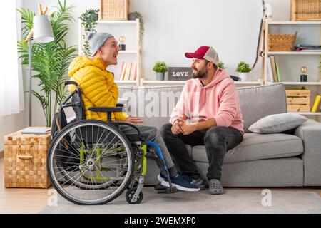 Uomo disabili in sedia a rotelle e amici che chiacchierano rilassati nel soggiorno della casa Foto Stock