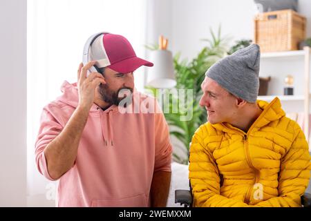 Uomo che ascolta musica con le cuffie accanto a un amico disabile in sedia a rotelle a casa Foto Stock