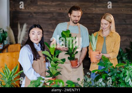 Gente in un negozio di fiori, cliente donna bionda matura che sceglie una pianta per la sua casa. Compratore che parla con due giardinieri in serra. Piccola impresa Foto Stock