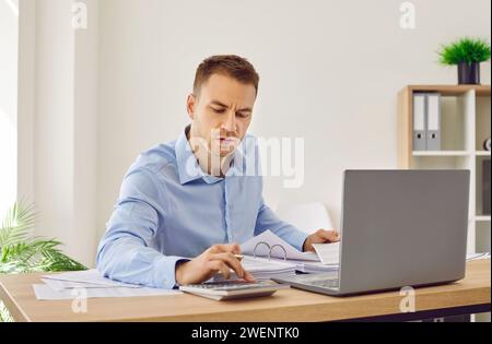 Commercialista di sesso maschile che lavora in ufficio, fa documenti cartacei e utilizza un computer portatile con calcolatrice Foto Stock