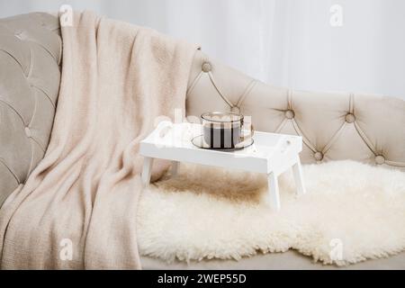 Vassoio per la colazione in legno bianco con le gambe sul divano di casa con una tazza di caffè caldo e candele in fiamme. Allestimento Hygge con coperta a quadri di lana morbida. Foto Stock