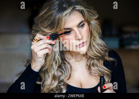 Donna elegante con ricci evidenziati applica meticolosamente il mascara, migliorando la sua bellezza naturale nel suo ambiente domestico. Foto Stock