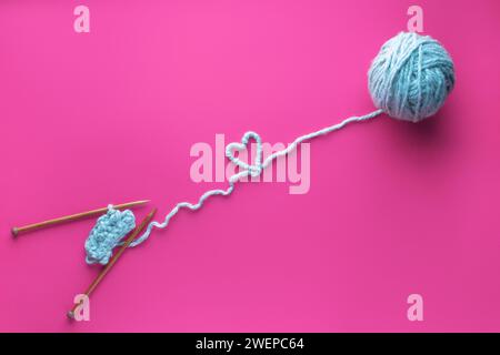 Sfera blu di filo e aghi per maglieria su sfondo rosa. Concetto di amore per la lavorazione del riccio e la lavorazione a maglia. Cuore fatto di fili. Foto Stock