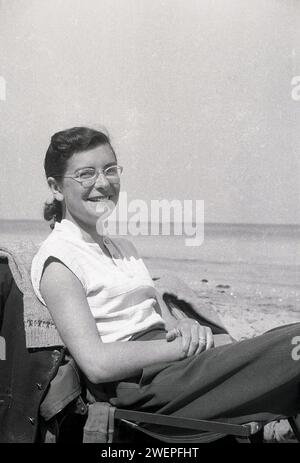 Anni '1960, storica, una signora in vacanza, rilassante sulla spiaggia di Margate, Inghilterra, Regno Unito, seduta su una sedia di metallo bassa, con sedile in tessuto sorridente davanti alla fotocamera. Foto Stock