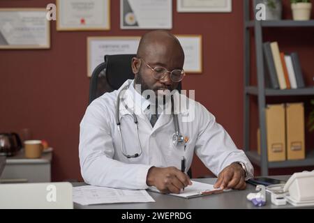 Medico afroamericano che scrive con attenzione su carta attaccata agli appunti seduto sul suo posto di lavoro Foto Stock