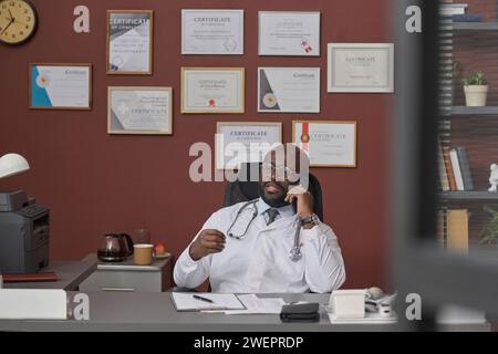 Medico afroamericano che parla sul suo smartphone mentre si trova al suo posto di lavoro in clinica Foto Stock