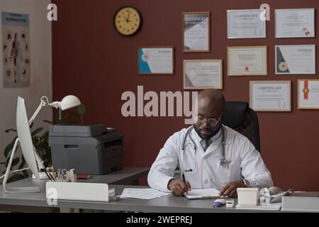 Un medico afroamericano seduto da solo alla scrivania del suo ufficio e che lavorava con i documenti Foto Stock