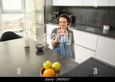 Ritratto di donna con volto sorridente e felice, che si gode il tè, chiude gli occhi dalla gioia, tiene una tazza calda in entrambe le mani, mangia cereali per Foto Stock