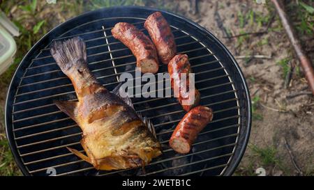 Vista dall'alto di pesce intero alla griglia fresco, spiedino, salsicce su griglia rotonda a carbone nero, sfondo di erba verde. Barbecue, grigliate e cibo Foto Stock