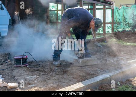 Un operaio edile taglia un marciapiede di calcestruzzo utilizzando una smerigliatrice angolare professionale causando una nuvola di polvere e fumo Foto Stock
