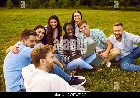 Amici universitari che lavorano e studiano insieme usando un computer portatile mentre sono seduti sull'erba del parco del campus. Foto Stock