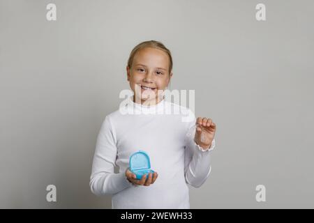 Simpatica bambina che tiene allineatori invisibili per sbiancare e raddrizzare i denti con una scatola blu colorata su sfondo bianco. Orthod Foto Stock