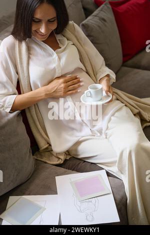 Una donna incinta sorridente è seduta sul divano con una tazza di caffè Foto Stock