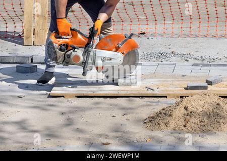 Un lavoratore taglia lastre di pavimentazione su un pallet di legno con una fresa manuale a gas con lama diamantata in una chiara giornata estiva. Copia spazio. Foto Stock