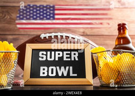 Germania - 27 gennaio 2024: Scritta del Super Bowl su una tavola circondata da spuntini, birra e football americano per il Super Bowl insieme a una bandiera degli Stati Uniti. Evento sportivo in America FOTOMONTAGGIO *** Super Bowl Schriftzug auf einer Tafel umgeben von Snacks, Bier und ein American Football zum Super-Bowl zusammen mit einer USA Fahne. Sportveranstaltung in Amerika FOTOMONTAGE Foto Stock