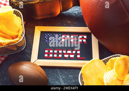 Germania - 27 gennaio 2024: Scritta del Super Bowl su una tavola circondata da spuntini, birra e football americano per il Super Bowl. Grande evento sportivo in America FOTOMONTAGGIO *** Super Bowl Schriftzug auf einer Tafel umgeben von Snacks, Bier und ein American Football zum Super-Bowl. Großveranstaltung im Sport in Amerika FOTOMONTAGE Foto Stock