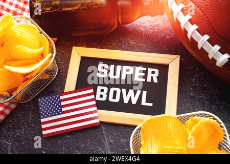 Germania - 27 gennaio 2024: Scritta del Super Bowl su una tavola circondata da spuntini, birra e football americano per il Super Bowl insieme a una bandiera degli Stati Uniti. Evento sportivo in America FOTOMONTAGGIO *** Super Bowl Schriftzug auf einer Tafel umgeben von Snacks, Bier und ein American Football zum Super-Bowl zusammen mit einer USA Fahne. Sportveranstaltung in Amerika FOTOMONTAGE Foto Stock