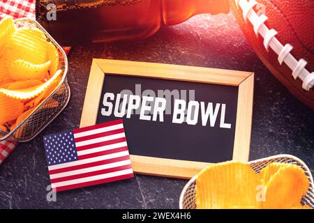 Germania - 27 gennaio 2024: Scritta del Super Bowl su una tavola circondata da spuntini, birra e football americano per il Super Bowl insieme a una bandiera degli Stati Uniti. Evento sportivo in America FOTOMONTAGGIO *** Super Bowl Schriftzug auf einer Tafel umgeben von Snacks, Bier und ein American Football zum Super-Bowl zusammen mit einer USA Fahne. Sportveranstaltung in Amerika FOTOMONTAGE Foto Stock