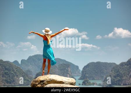 Bella giovane donna in abito blu e cappello bianco è in piedi a braccia aperte su una grande roccia con vista sul mare e sulle isole. Provincia di Phang Nga, Thailandia Foto Stock