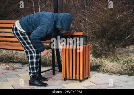 Un vecchio uomo caucasico senzatetto ruba per cibo e spazzatura in un cestino in autunno in un parco Foto Stock