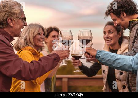 Gruppo di amici sorridenti che brindano con bicchieri di vino rosso all'aperto, celebrando la gioia della compagnia al tramonto. Foto Stock