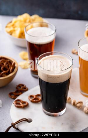 Birra scura, birra lager e birra ale in bicchieri alti sul tavolo, varietà di birra con spuntini, pretzel e patatine fritte Foto Stock