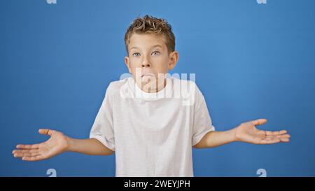 Un ragazzo biondo, con un'espressione adorabile, incerto su un isolato sfondo blu, il suo dubbio aggiungeva un tocco di delicatezza alla sua confusione. dubitare, lui Foto Stock