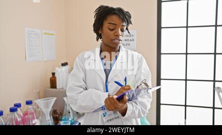 Scienziata africana che scrive appunti in un laboratorio con oggetti in vetro e istruzioni di sicurezza visibili. Foto Stock