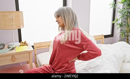 Donna ispanica matura con lunghi capelli grigi in un pigiama rosa si siede con cura su un letto in una camera da letto ben illuminata. Foto Stock