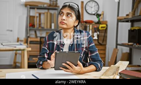Una donna indiana contemplativa con i capelli lunghi tiene una tavoletta in un laboratorio di lavorazione del legno, circondata da attrezzi e legno. Foto Stock
