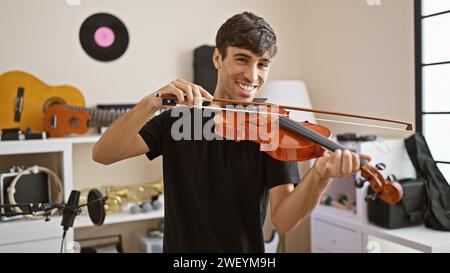 Giovane artista ispanico sicuro e sorridente che suona il violino felicemente in uno studio di musica al coperto, una sinfonia di intrattenimento classico Foto Stock