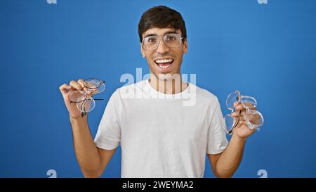 Allegro giovane ispanico, che sorride con sicurezza tenendo gli occhiali, in piedi isolato su uno sfondo blu freddo Foto Stock