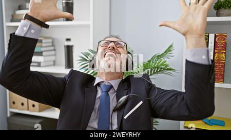 Uomo di mezza età sicuro di sé con i capelli grigi, un lavoratore professionale sorridente che indossa le cuffie, che celebra la sua vittoria in ufficio: Un vincitore nella sua i professionale Foto Stock