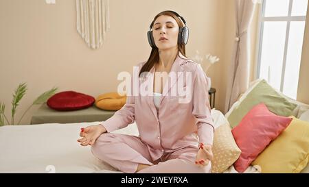 Una giovane donna tranquilla in pigiama rosa che medita su un letto al chiuso mentre ascolta musica con le cuffie. Foto Stock