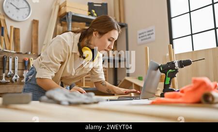 Donna concentrata che utilizza un computer portatile in un laboratorio di falegnameria con utensili e legno, incarnando un'atmosfera artigianale capace. Foto Stock