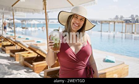 Una donna sorridente con cappello e costume da bagno gode di un mojito in una lussuosa piscina con vista mare Foto Stock