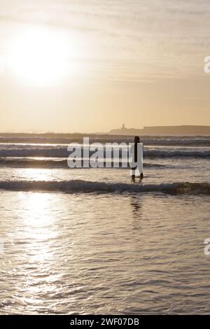 Man in silhouette si erge in acque poco profonde guardando verso il tramonto con l'isola alle spalle mentre le onde si infrangono a Essaouira, Marocco, 27 gennaio 2024 Foto Stock