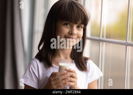 La felice preadolescente sta in piedi vicino alla finestra regge un bicchiere di latte Foto Stock