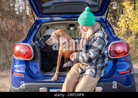 Donna proprietaria di animali domestici in viaggio con il cane magyar vizsla al guinzaglio seduto nel bagagliaio dell'auto sulla natura. Foto Stock